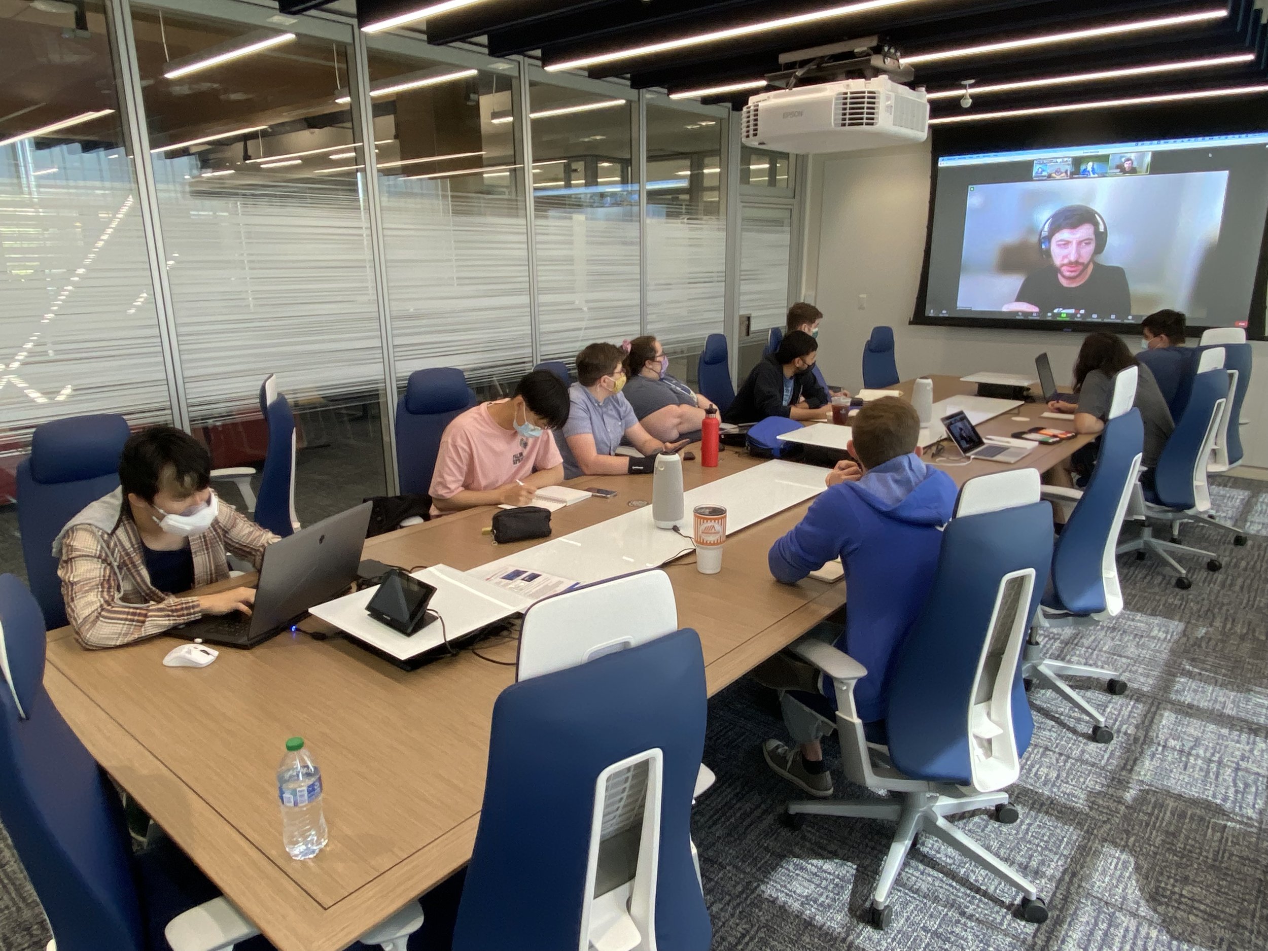 Photo of developers in a conference room. Projected onto the wall in front of them is a zoom meeting with a panel of experts.