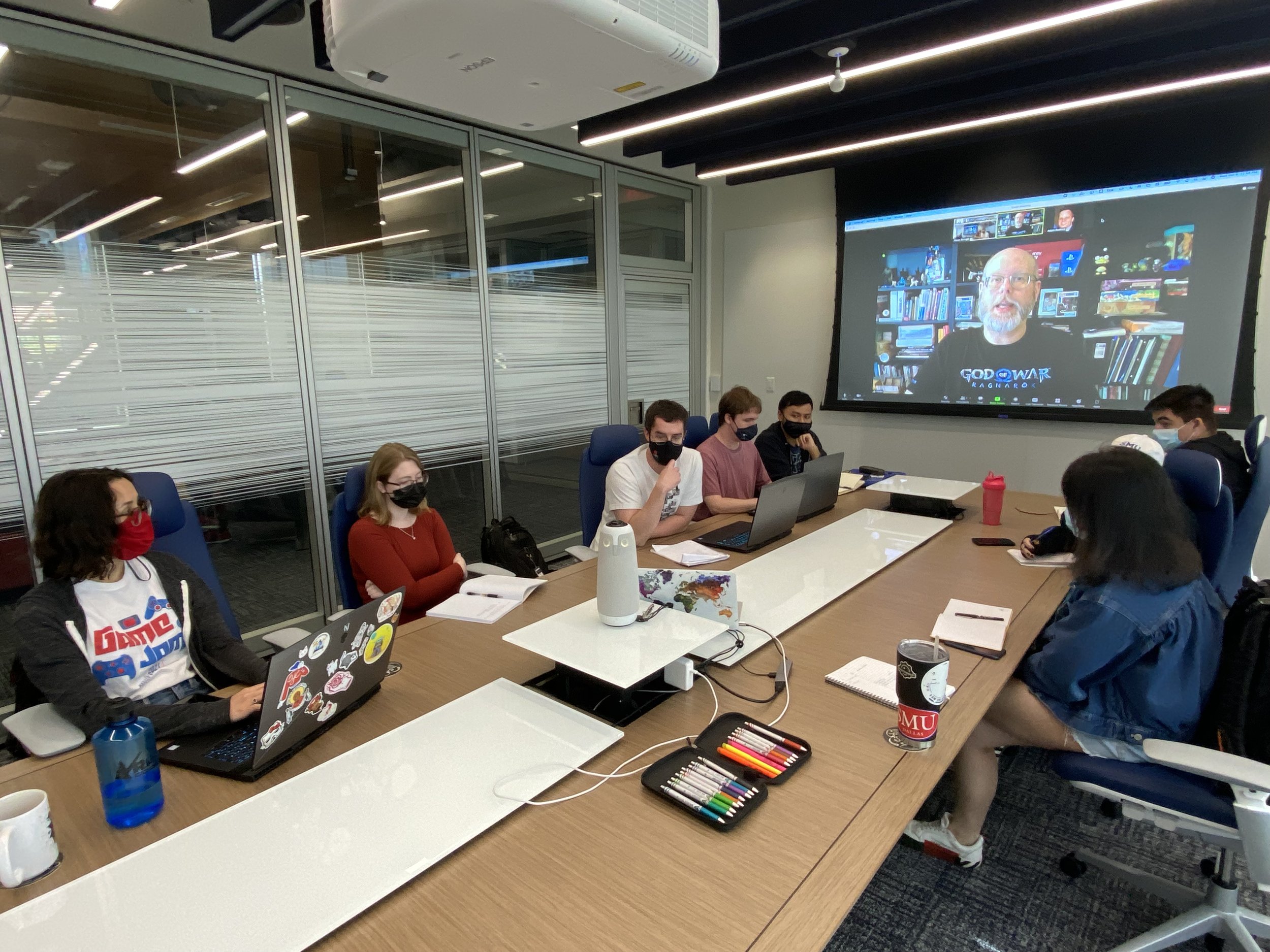 Photo of a group of Guildhall developers in a conference room. Projected onto the wall is a Zoom with a panel of experts giving the developers feedback.