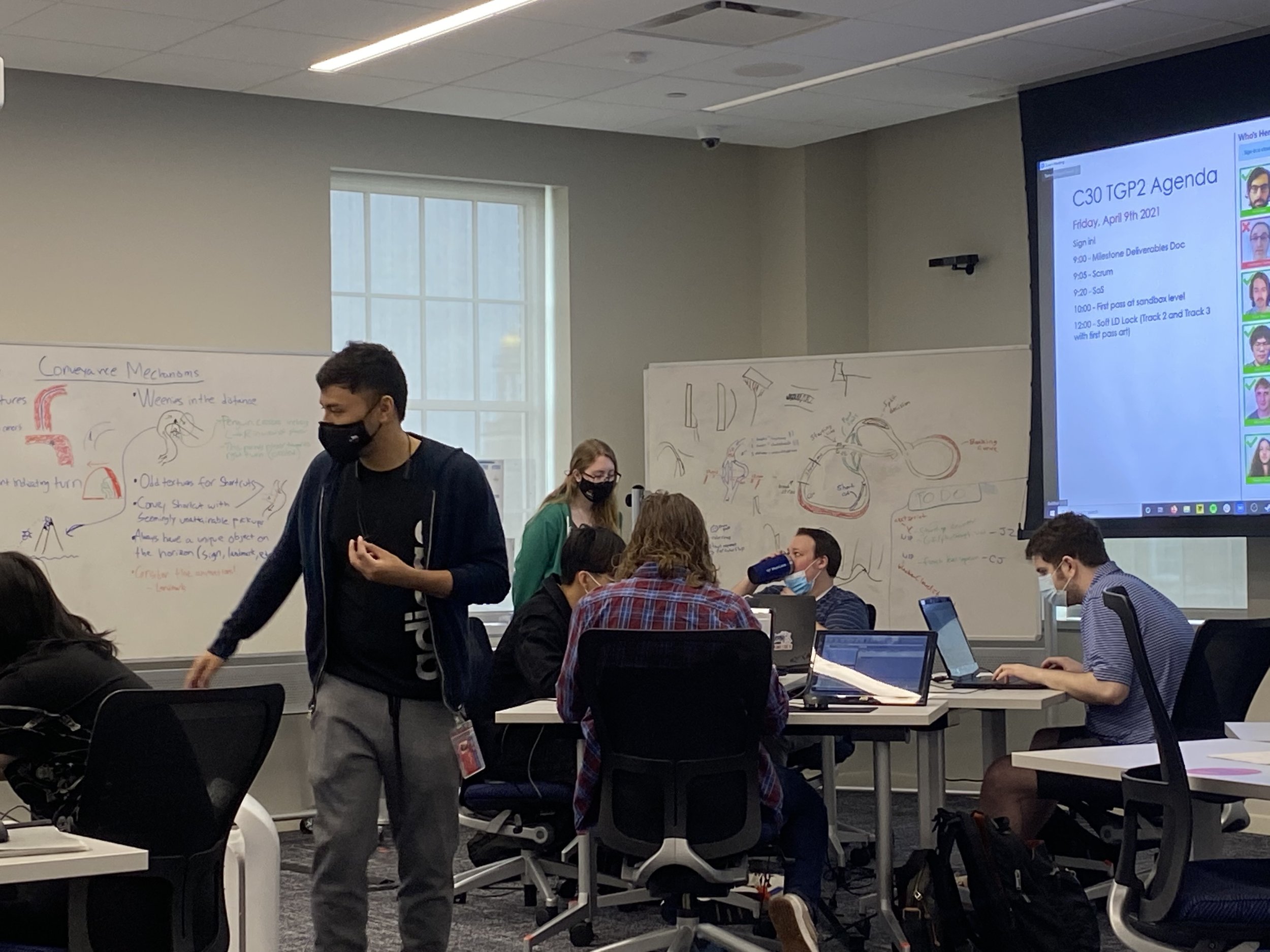 Photograph of the level designer's section of the room. In the foreground, Will walks around a desk. In the background, Dube speaks with a designer.