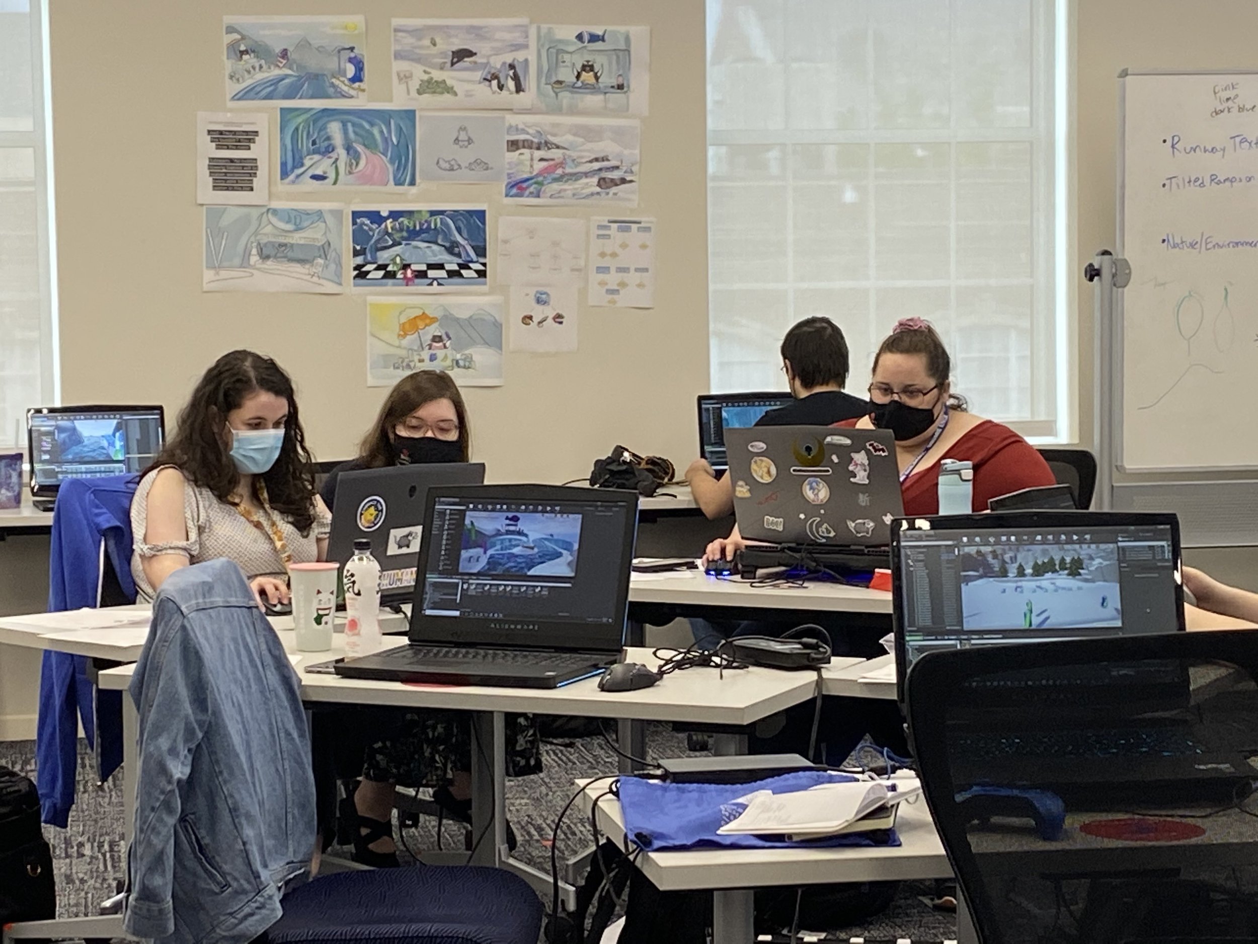 Photograph of the level designer's section of the room. Several designers can be seen working at their computers. On the wall behind them is a collage of concept art for Snowpainters.