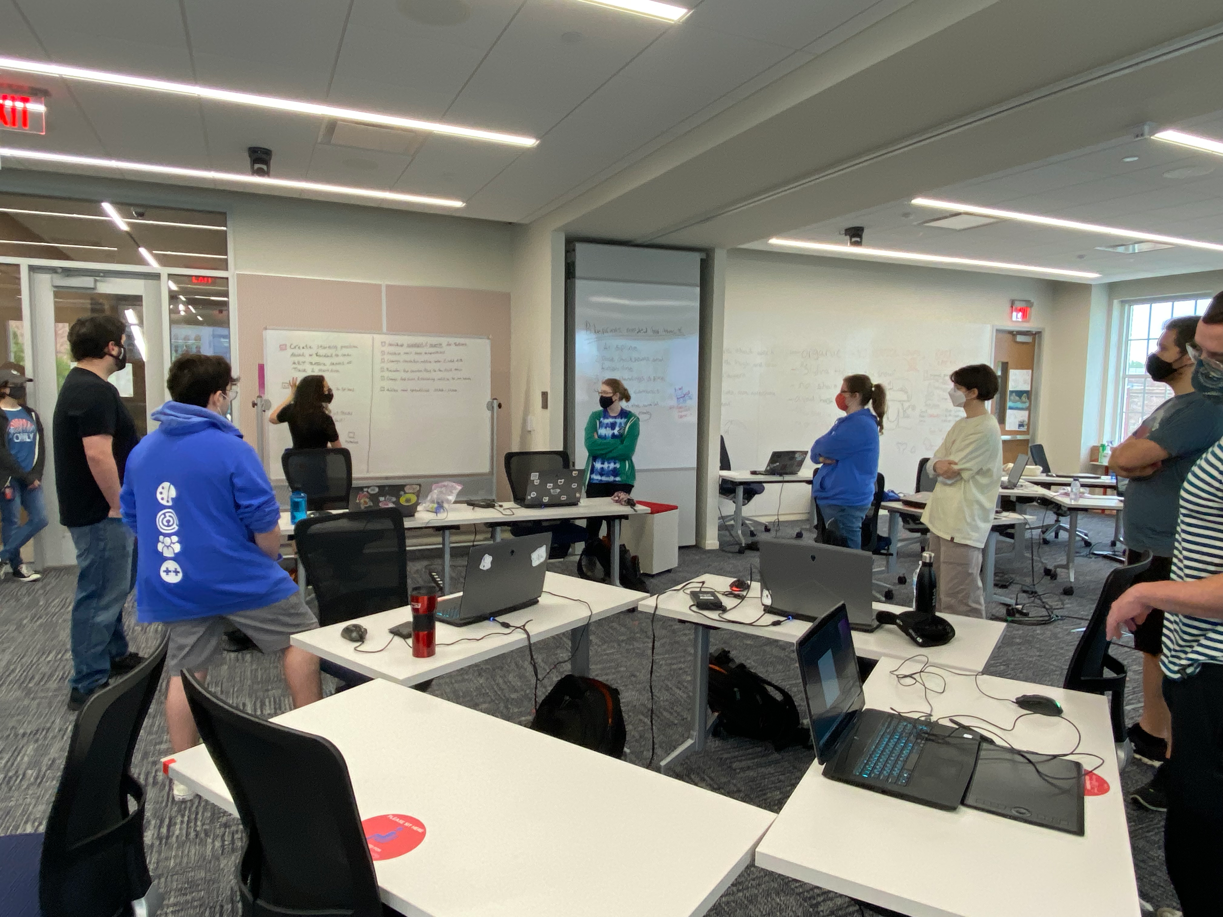 Photograph of a group of people standing in front of their desks. Nooka stands in front of the whiteboard, writing down people's tasks.